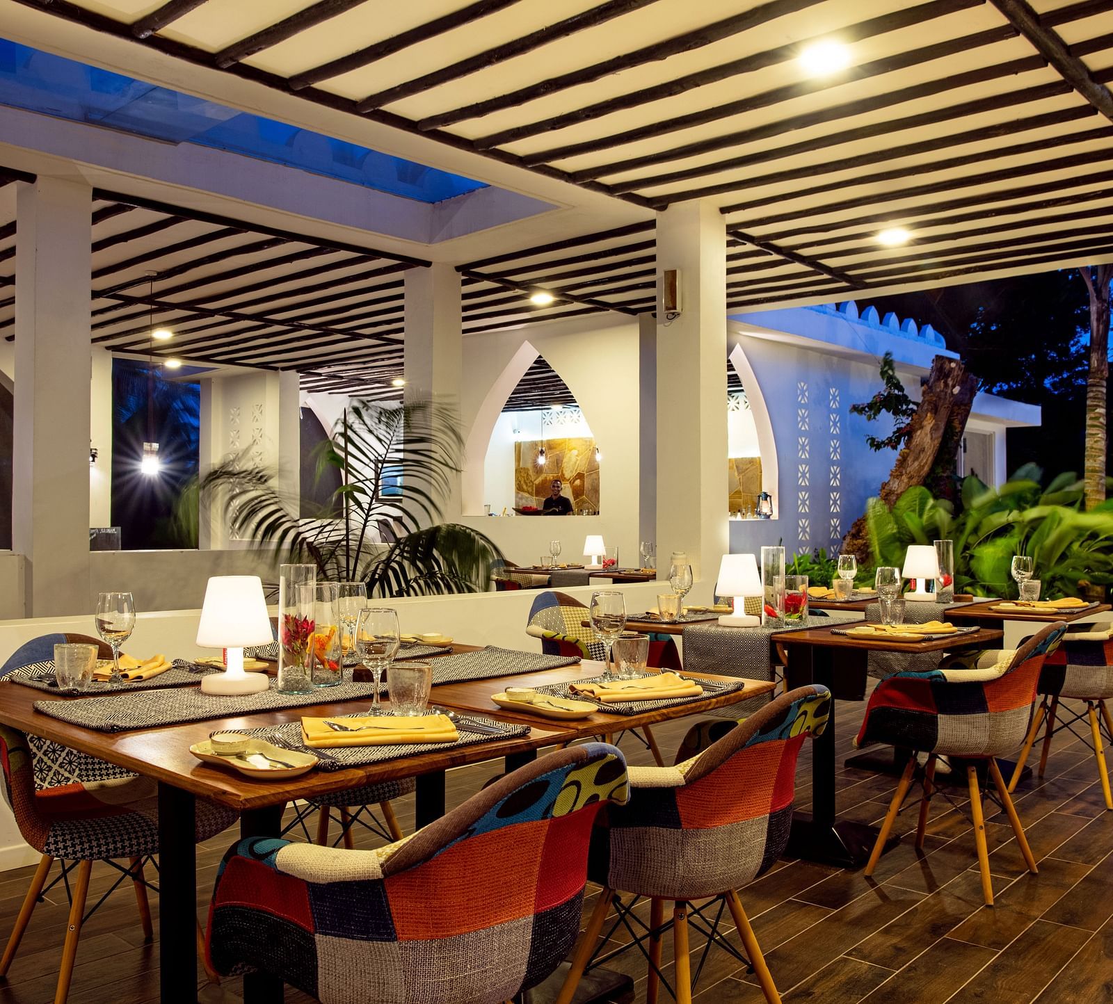 A well-lit indoor restaurant dining area with wooden tables set with glasses, plates, and napkins, surrounded by patterned chairs and decorative lighting at Shooting Star boutique hotel.