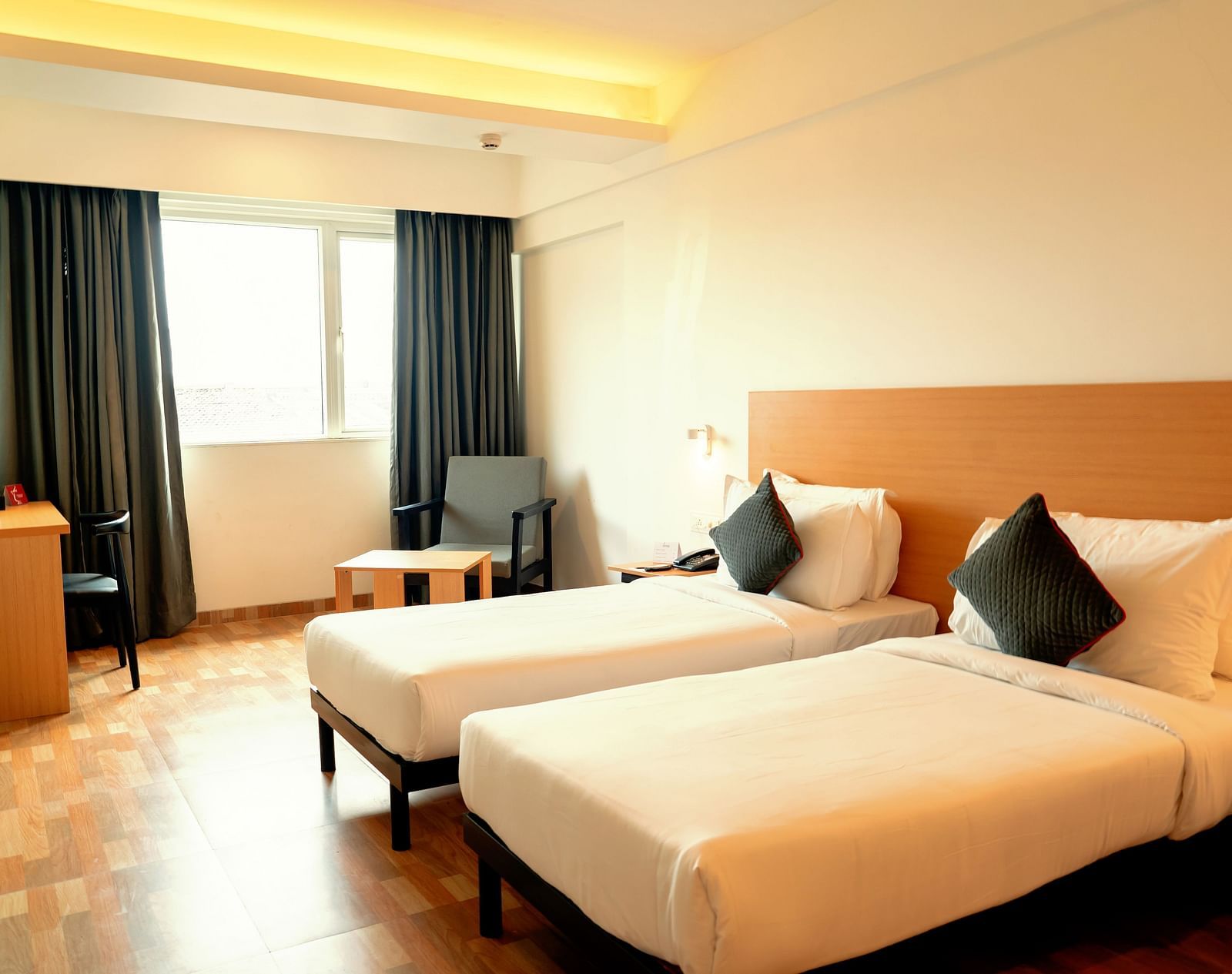 A bright hotel room with two single beds, a desk and a flat-screen TV, with a large window draped in gray curtains - TGI Ameya Grand, Goa