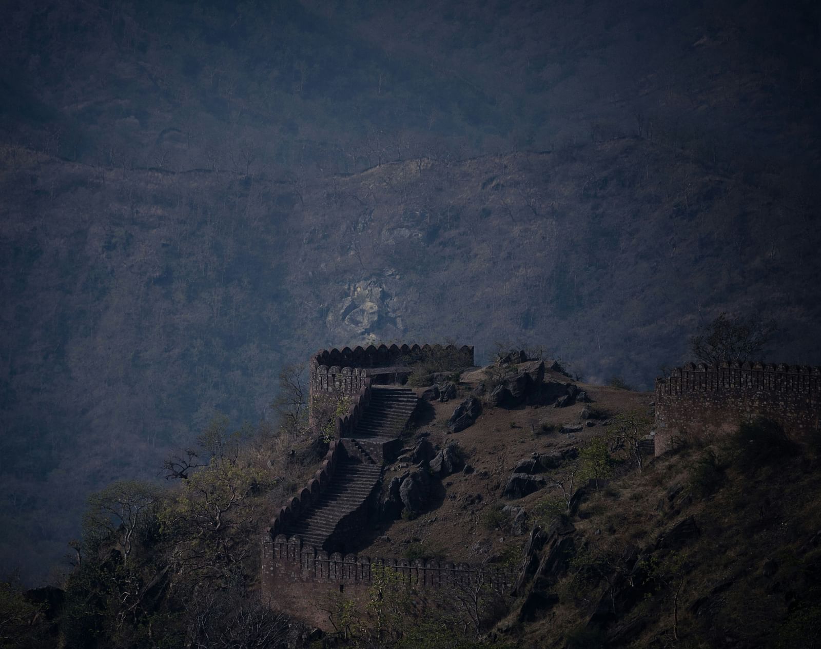 A weathered stone fort wall with zig-zag stairs and battlements on a rocky hill, against a hazy mountain backdrop