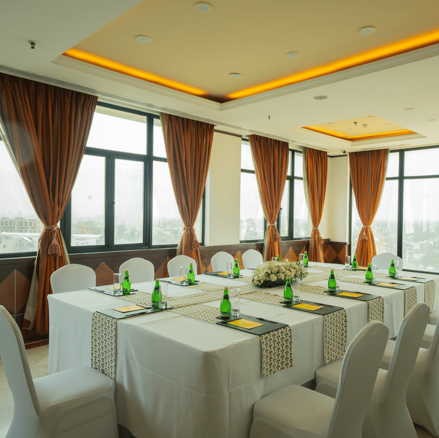 Sampan Tea Room at The Suryaa set with rectangular tables, white covers, green bottles, notepads, pens, warm recessed lighting, large windows with city view and brown curtains.