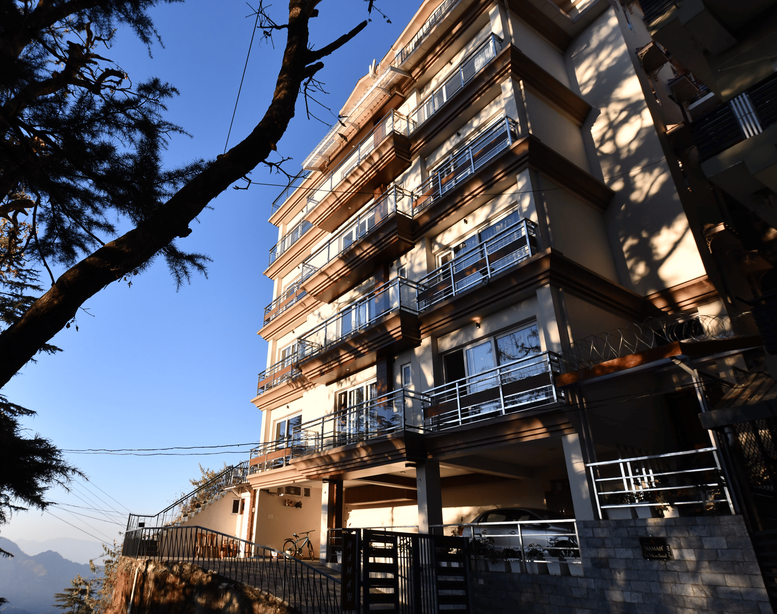 A facade of Perfectstayz Value Shimla (Namah Retreat) with balconies viewed from below against a clear sky and a tree in foreground.