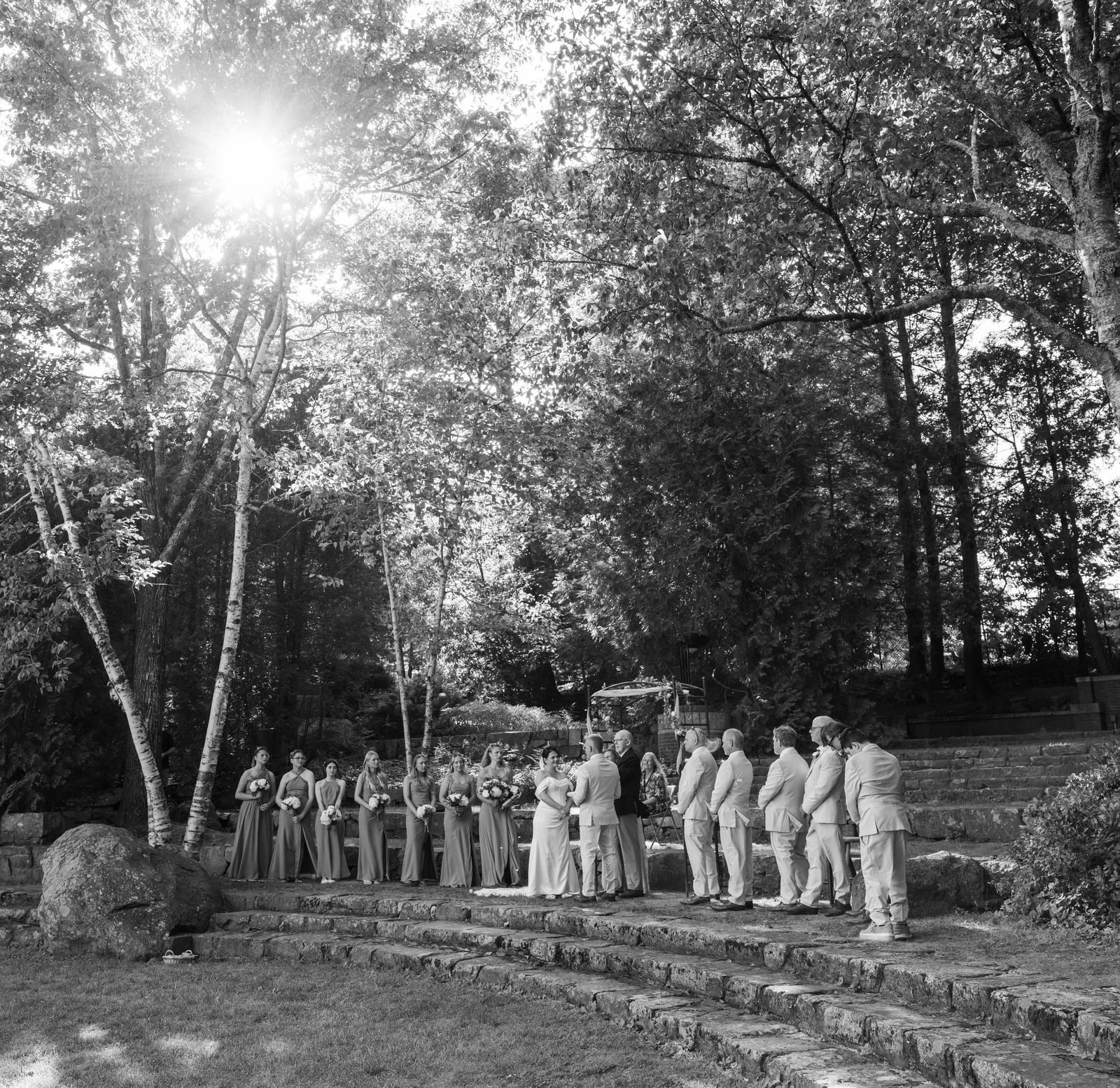 Wedding ceremony at Hartstone Inn with the couple, officiant, bridesmaids, and groomsmen standing on stone steps surrounded by trees and sunlight filtering through.