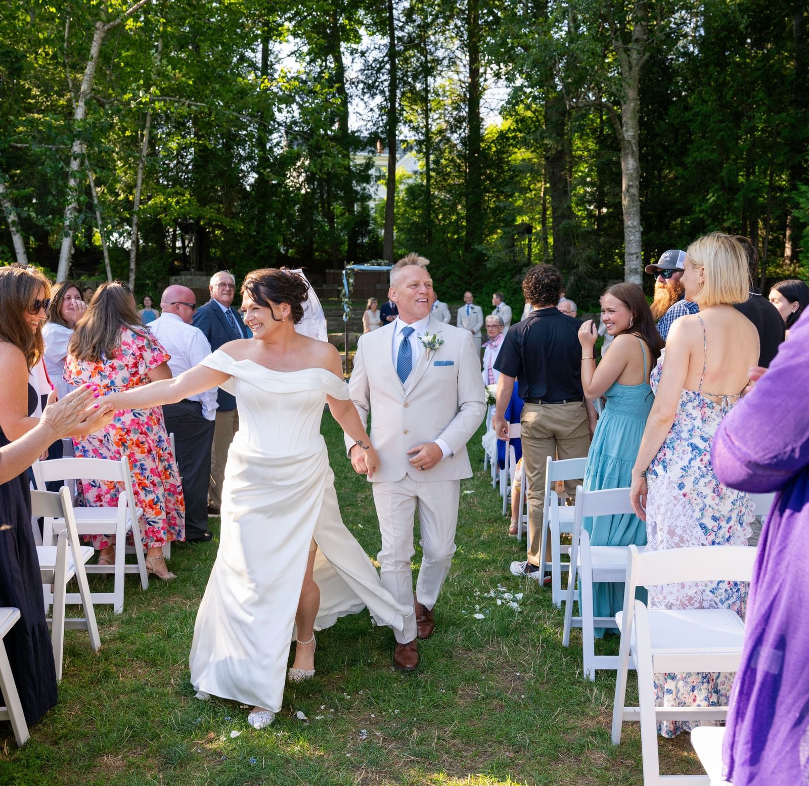 At Hartstone Inn, a newlywed couple walks down the aisle holding hands, surrounded by standing guests who are smiling, clapping, and reaching out to greet them.