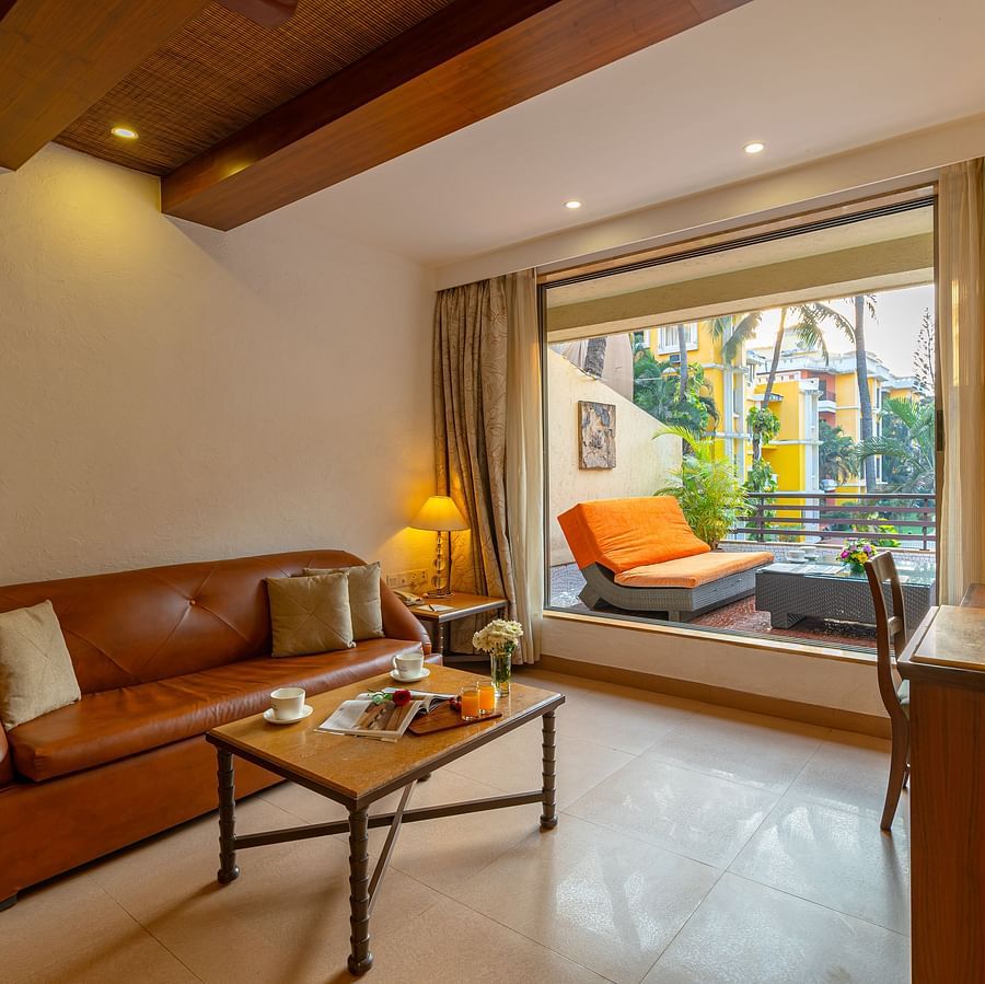 An image of a living room with a brown sofa, coffee table, table lamps, a tea and coffee maker, and an attached balcony with seating.