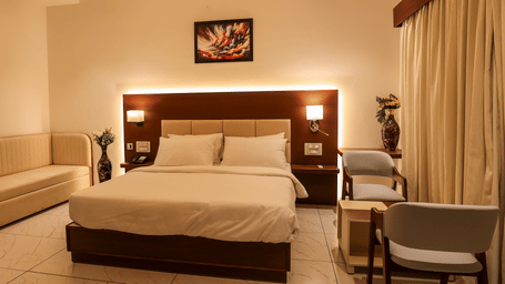 A well-lit hotel bedroom featuring a double bed, a beige sofa against the wall, an illuminated headboard, and a desk chair | Lords Sahyadri Resort, Shivamogga