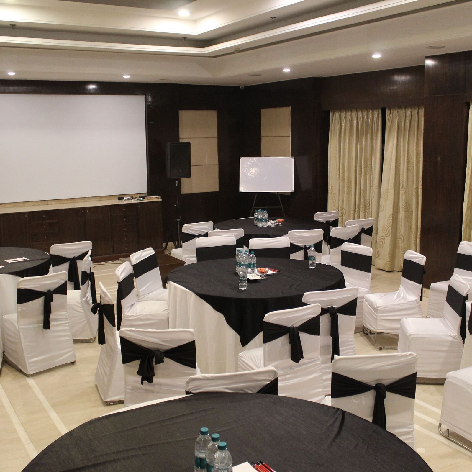 An image of a banquet hall at Inde Hotel Signature Tower, Gurgaon featuring a few round tables, each surrounded by chairs