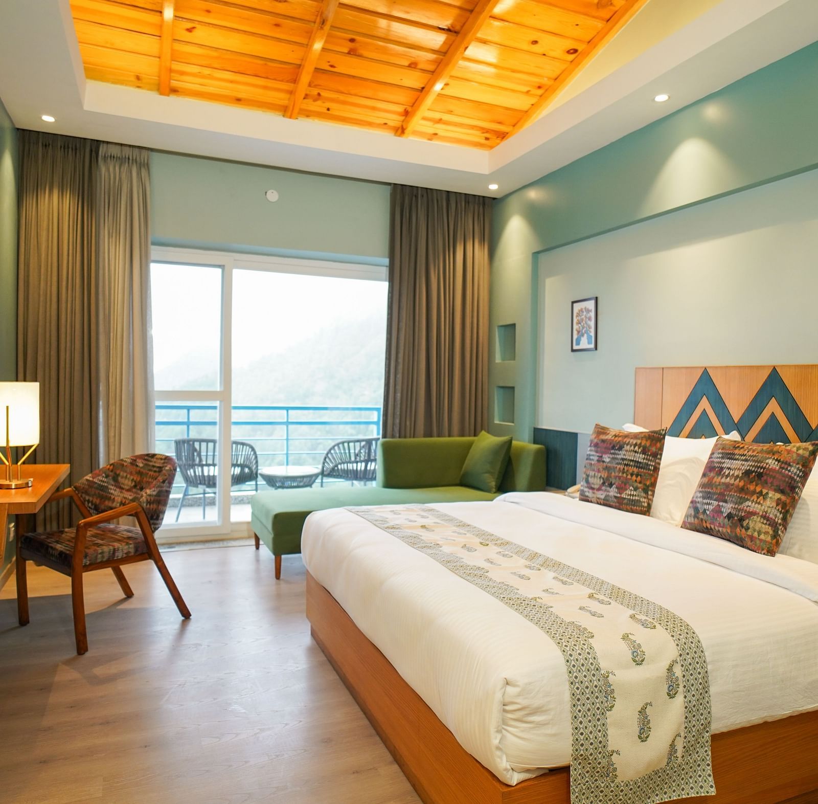 A spacious hotel room includes a large bed, a comfortable seating area with a sofa, and a balcony view of the hills | Deluxe Room with Valley View | Sukama Resort & Spa, Nainital