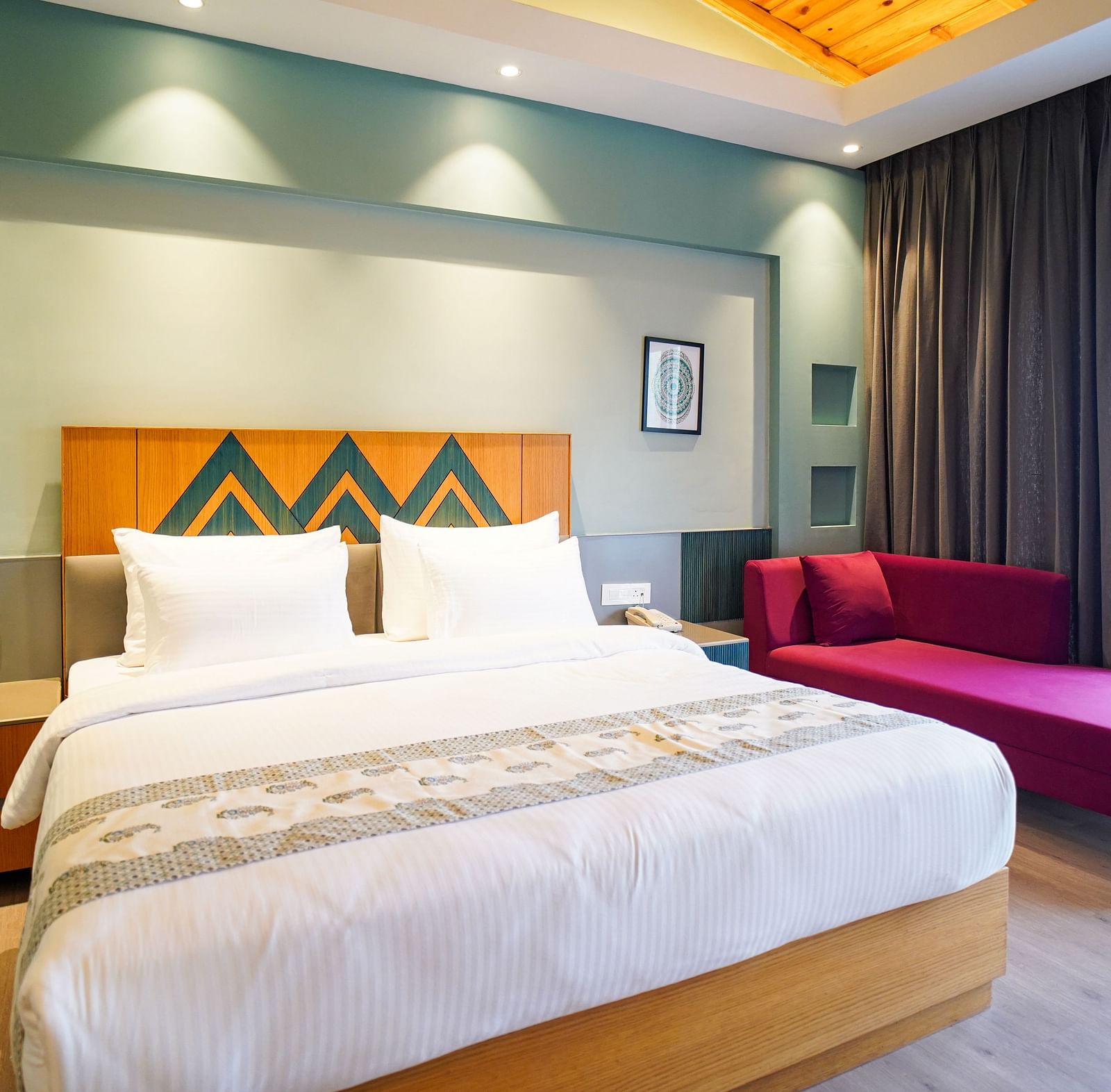 A hotel room displays a neatly made bed with white linens and a wooden headboard, accompanied by a purple chaise lounge and floor-to-ceiling curtains | Deluxe Room with Valley View | Sukama Resort & Spa, Nainital
