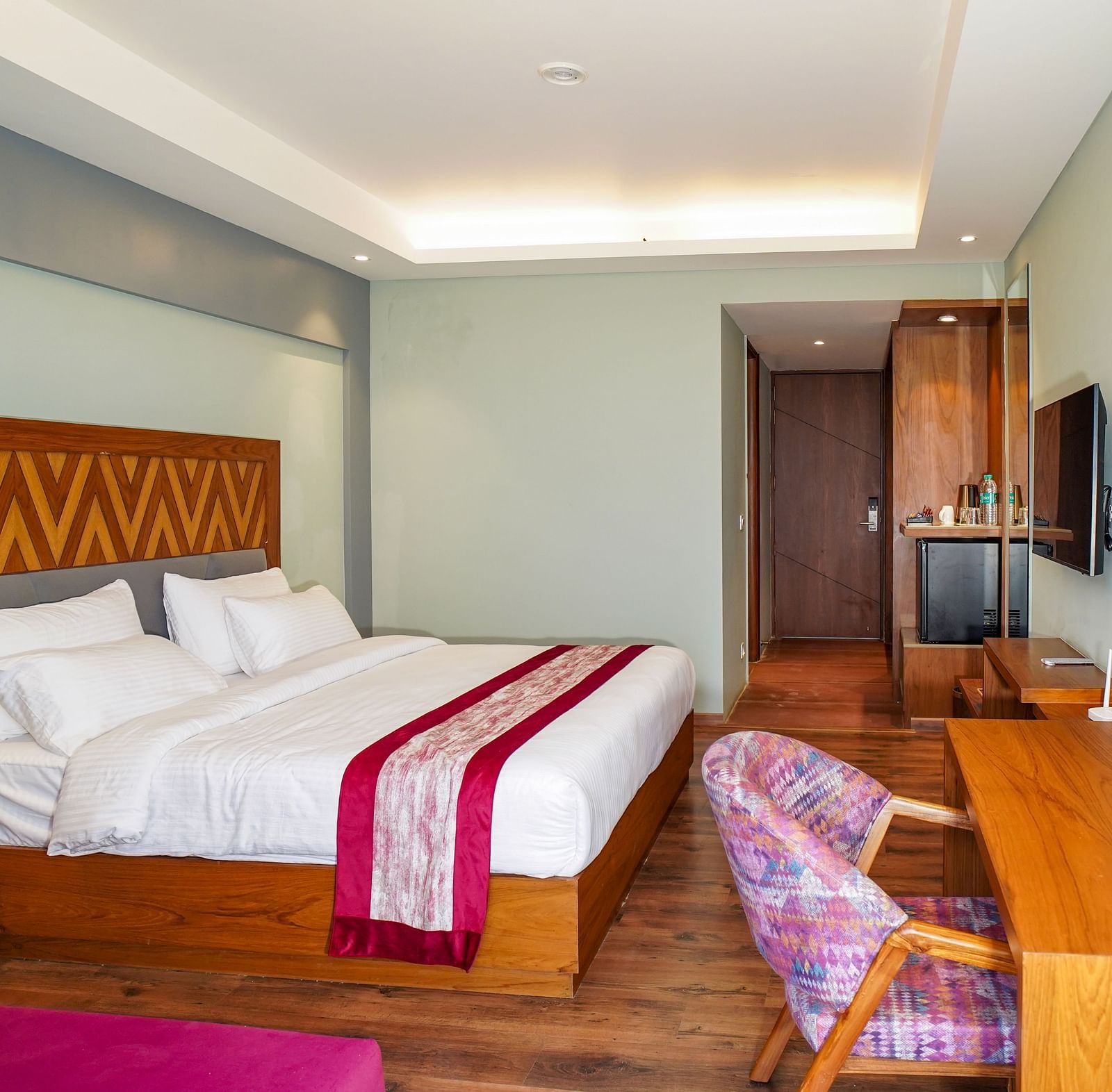 A spacious hotel bedroom is furnished with a large bed, a desk area, and a television, all in warm tones | Deluxe Room with Himalayan View | Sukama Resort & Spa, Nainital