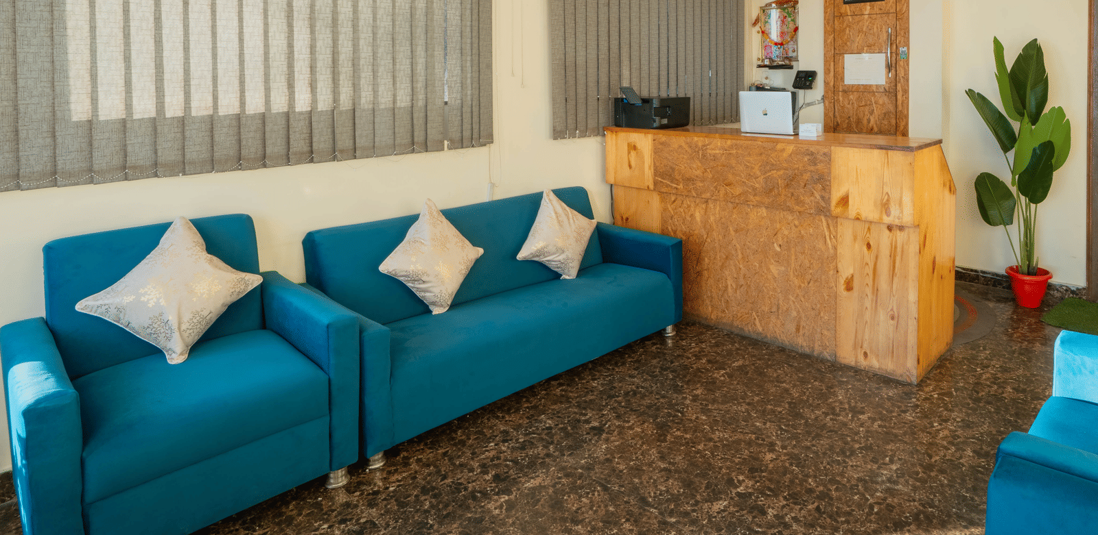A hotel lobby or waiting area, featuring teal blue sofas and a reception desk in the background, with vertical blinds on the windows | Perfectstayz Value Valley Vista