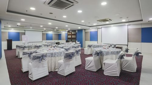 A spacious and elegantly designed conference hall with round tables and chairs draped in white covers, featuring advanced lighting and projection equipment.