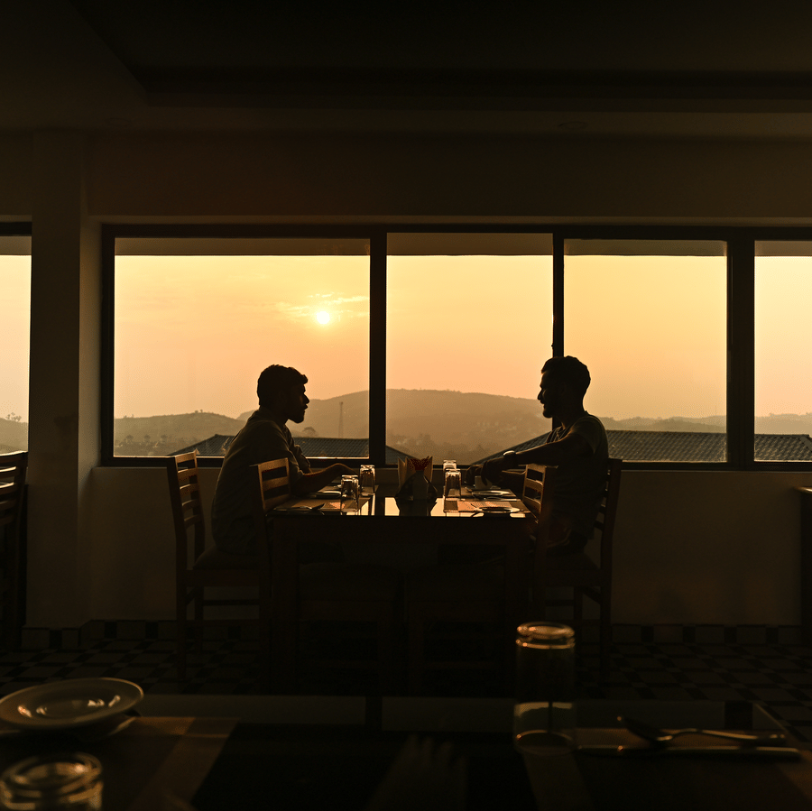 Two people sit at a dining table silhouetted against a large window showing a sunset over hilly terrain at one of the best restaurants in Vagamon at Abad Silvermist, Vagamon