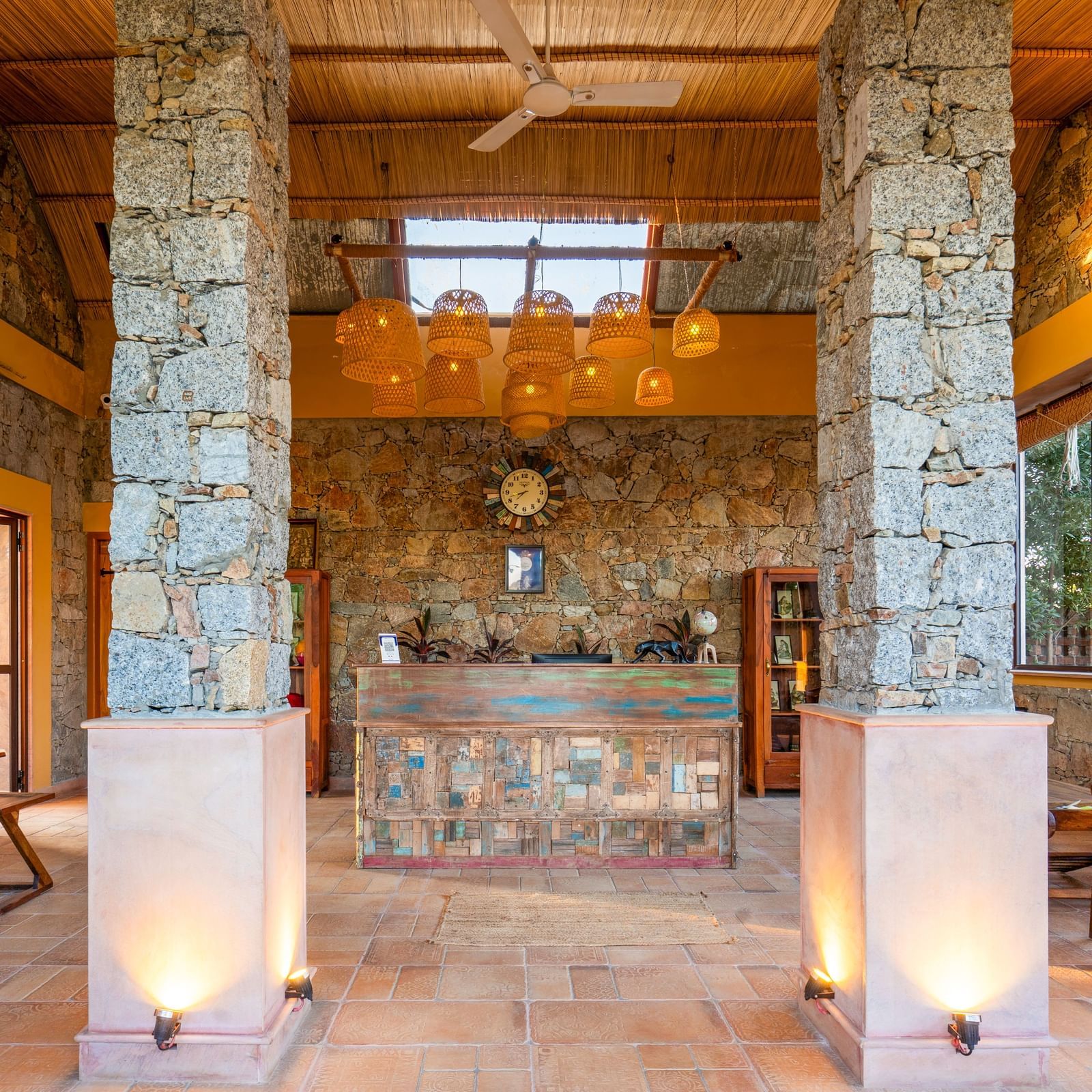 An image of a reception in bohemian theme and rock pillars and walls featuring  wooden seating area and a reception desk, and above it there are bamboo lamps - Kurja Jawai