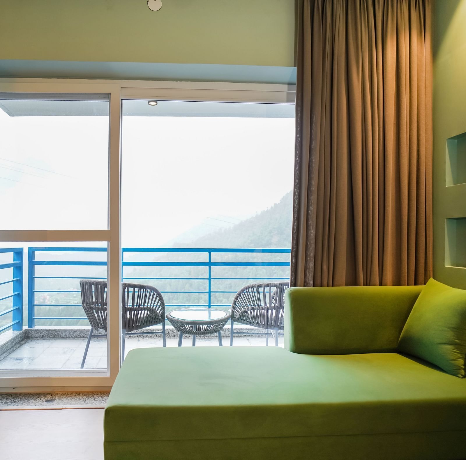 A modern hotel room with a green sofa and a large sliding glass door opening to a balcony with outdoor seating and a view of the hills | Deluxe Room with Valley View | Sukama Resort & Spa, Nainital