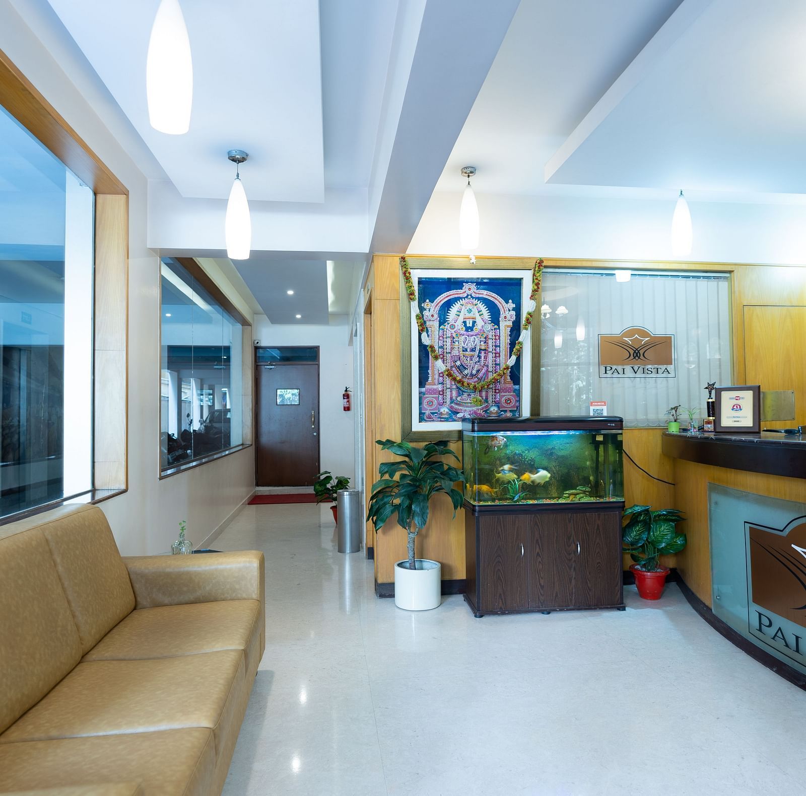 An image of a reception lobby featuring a reception desk, a sofa opposite to it, and an aquarium on one side of the room - Hotel Pai Vista, Mysore