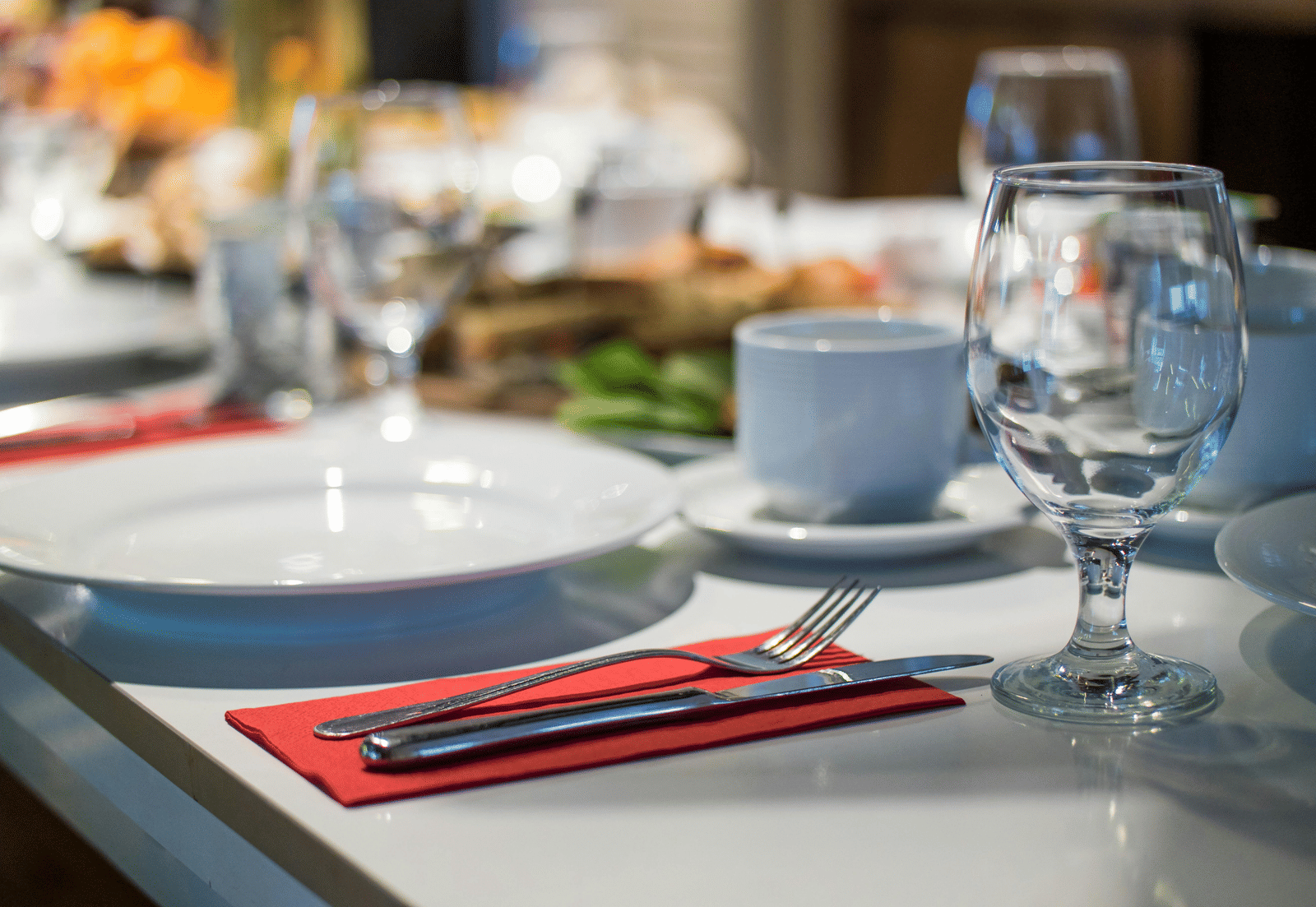A close-up, blurred shot of a formal dinner table setting with white plates, red napkins, silverware, and clear wine glasses.