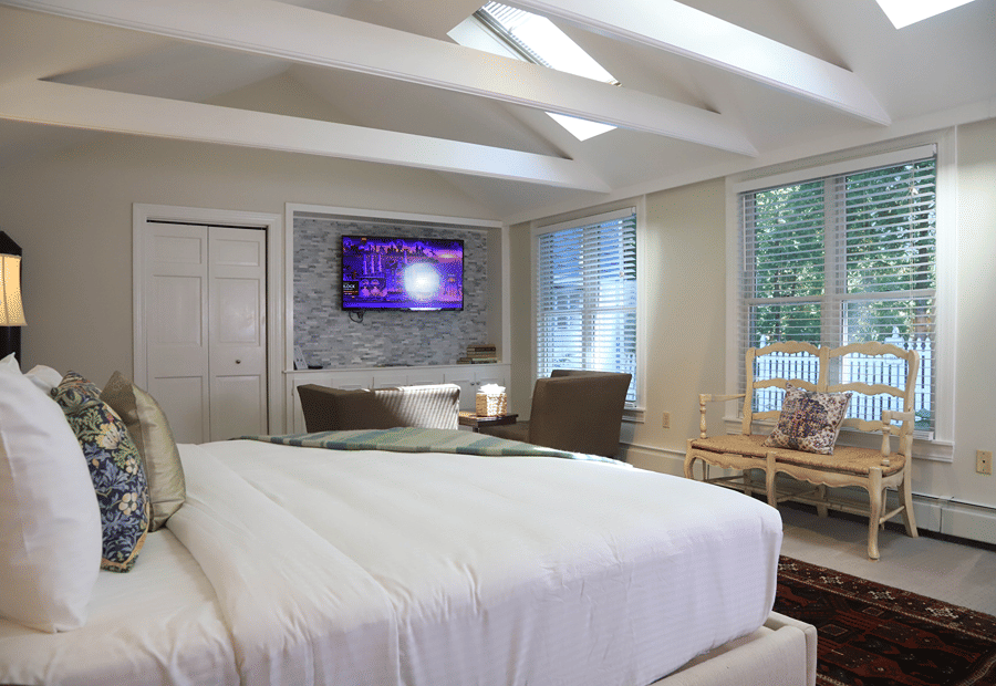 A wide-angle view of a guest room with a large bed, a TV mounted on a stone accent wall, and a window with a wooden bench underneath.