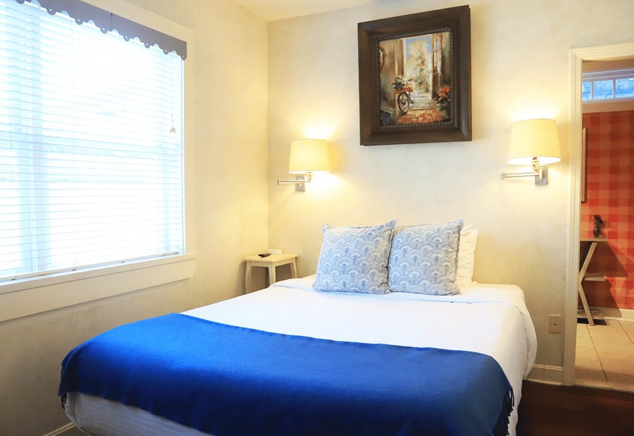 A neatly made bed with a blue spread and white pillows in a room with white walls, a window, and a painting above the bed.