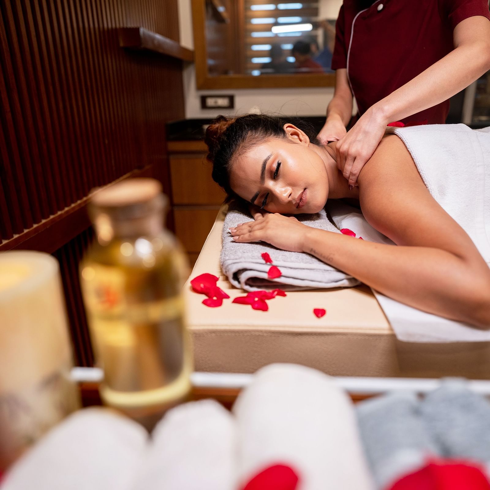 A lady lies on the bed, receiving a massage, with essential oils and rolled towels in the foreground