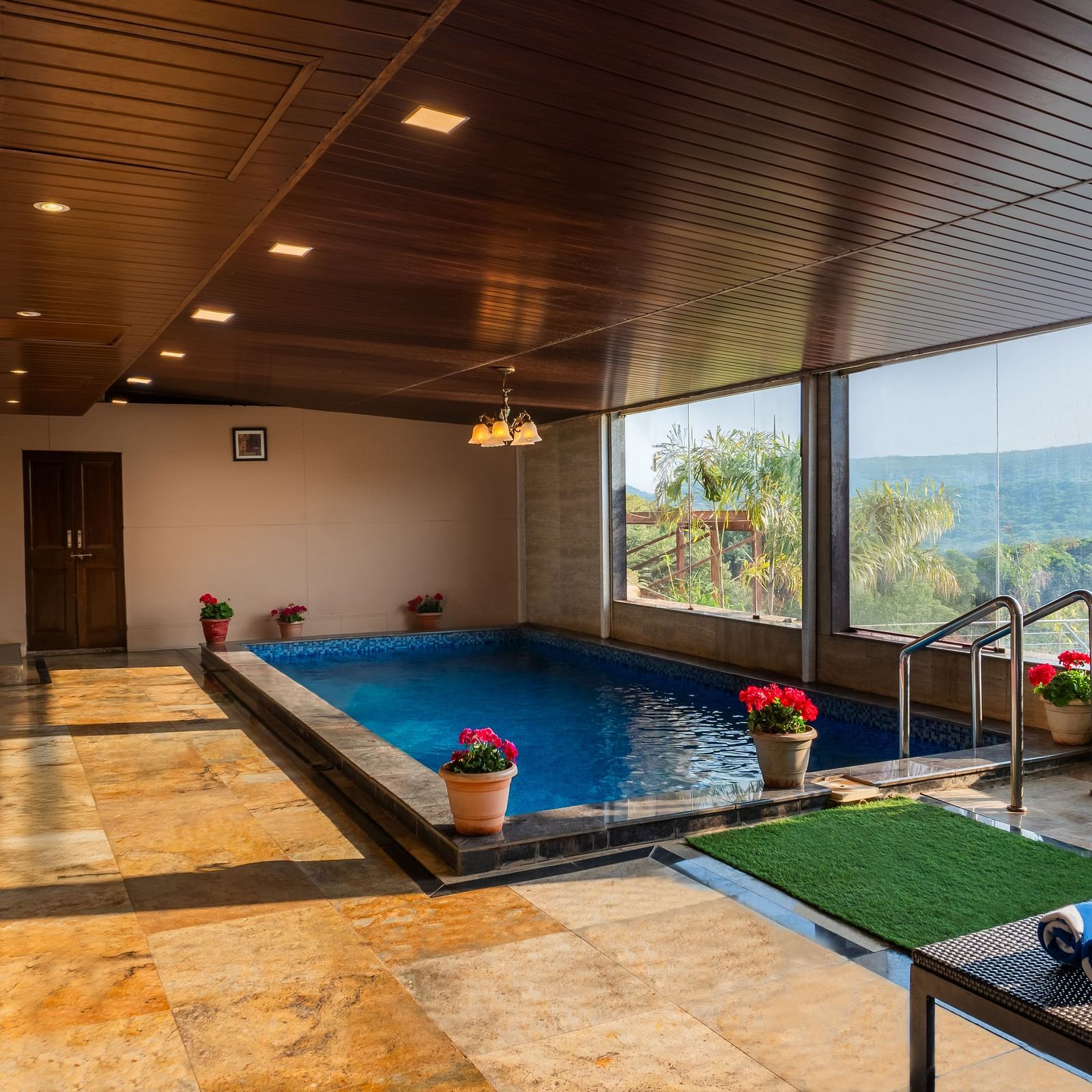 A bright indoor pool reflects the wood ceiling and recessed lights. Large windows reveal a scenic outdoor view. Lounge chairs with blue striped towels sit poolside on a stone floor next to potted flowers and a palm - The Grand Legacy Resort and Spa