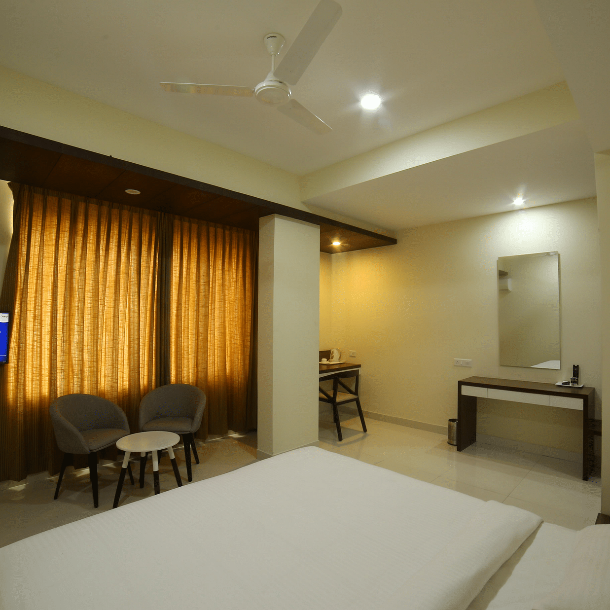 A room featuring a neat bed a smart TV a vanity area sofa chairs and a table - Mysore Gate Hotel Bangalore