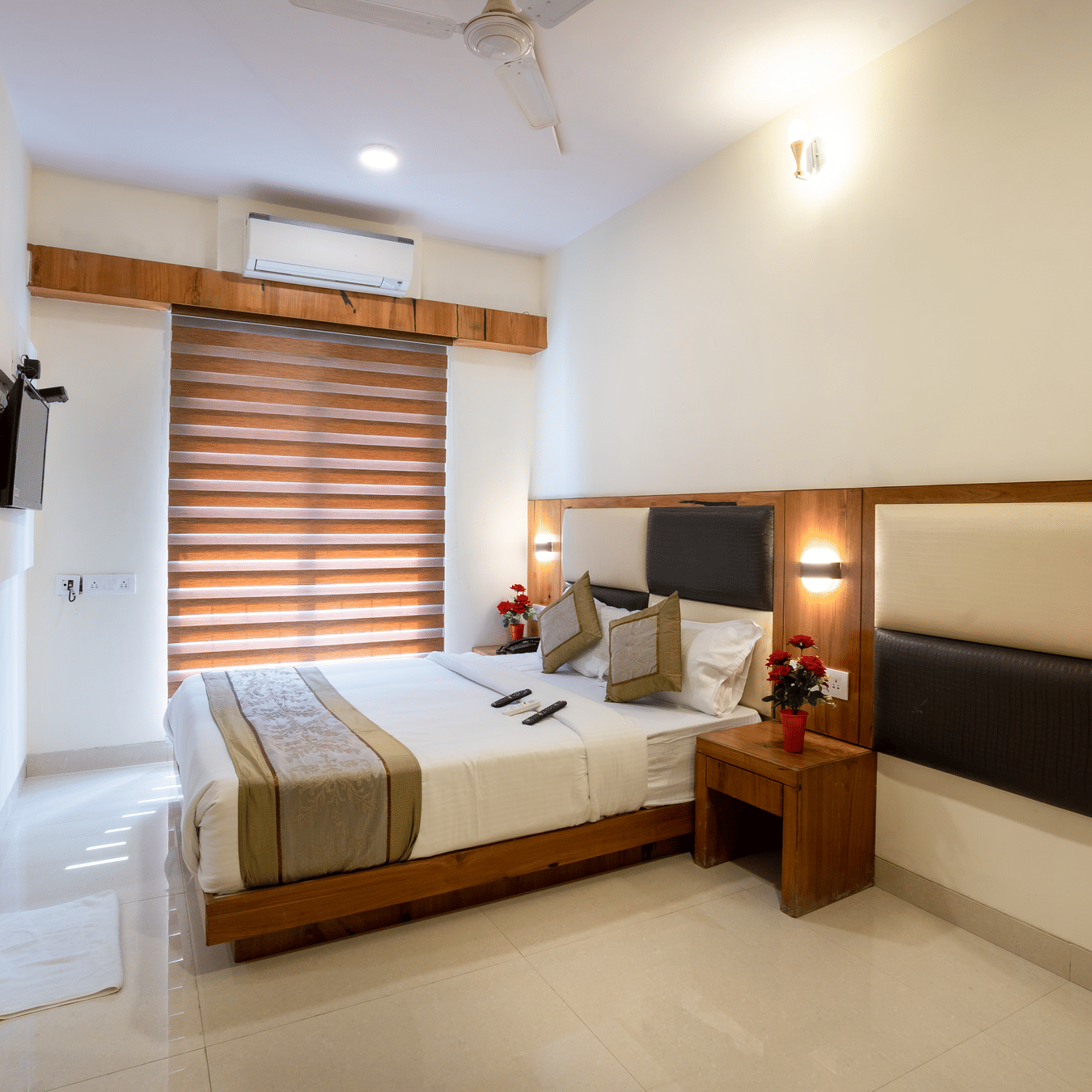 A bedroom at Home2 India with a double bed, pillows, bedside table, wall lights, window with blinds, wall paneling, and a door visible on one side
