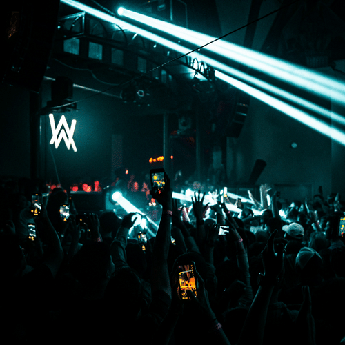  A packed nightclub with a large crowd facing a stage with a prominent AW logo. Multiple spotlights illuminate the scene, and the audience is holding up their phones, capturing the moment.
