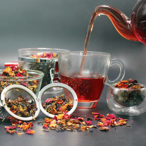 A teapot pours a reddish liquid into a clear mug, surrounded by loose tea leaves, dried flowers, and two metal tea infusers.
