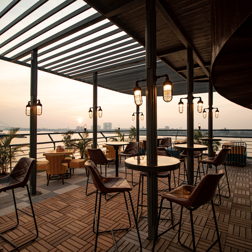 A rooftop dining area with wood flooring, modern furniture, and pendant lights at dusk, overlooking a suspension bridge and river.