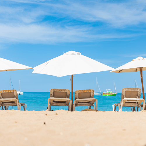 A row of empty loungers under white umbrellas lines a serene beachfront at The Soco Hotels, facing the clear blue sky and calm ocean.