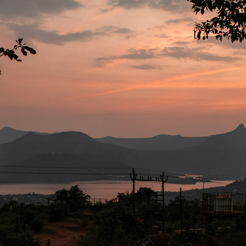 An overview of Pawna Lake in Lonavala, as seen during twilight hour with orangish hues in the sky and mountains in the background.