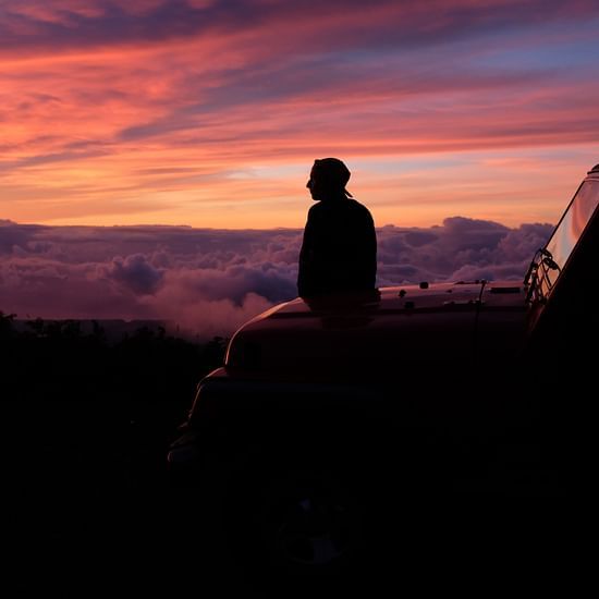 a person sitting on the hood of a car looking at the different hues in the sky