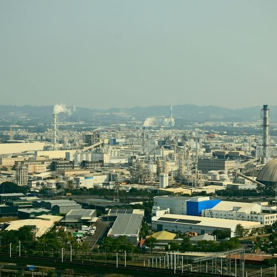 A wide-angle shot of a factory complex on a sunny day. In the foreground, there are green bushes. Behind them, two large blue shipping containers are stacked on an elevated platform, connected to a conveyor belt system. In the background are several indust