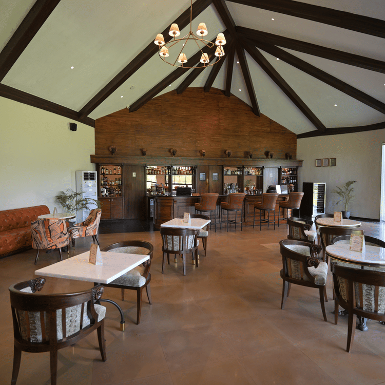 The Sariska Lounge of Ananta Spa & Resort Ajabgarh featuring a spacious seating area with wooden flooring, round tables, upholstered chairs, high vaulted ceiling with exposed beams, and a central chandelier creating a warm and inviting ambience.