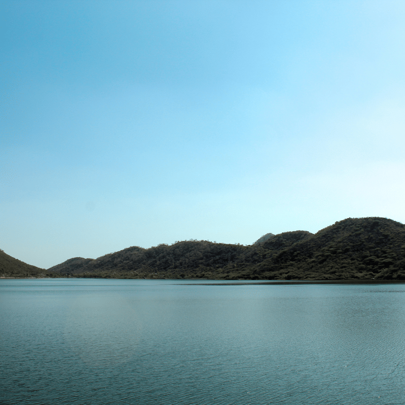 Wide view of calm lake water with low forested hills and a clear blue sky in the background