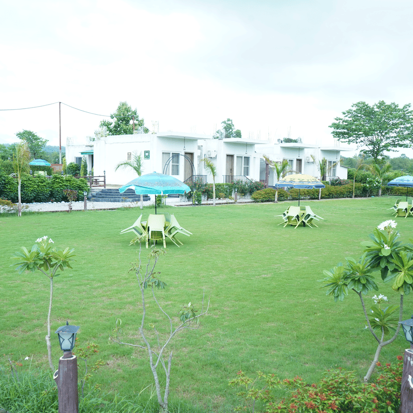 Scenic wide view of single-story resort cottages nestled behind a spacious green garden lawn adorned with small trees and outdoor table seating - Five Elements Hotels, Corbett Crown Resort, Jim Corbett
