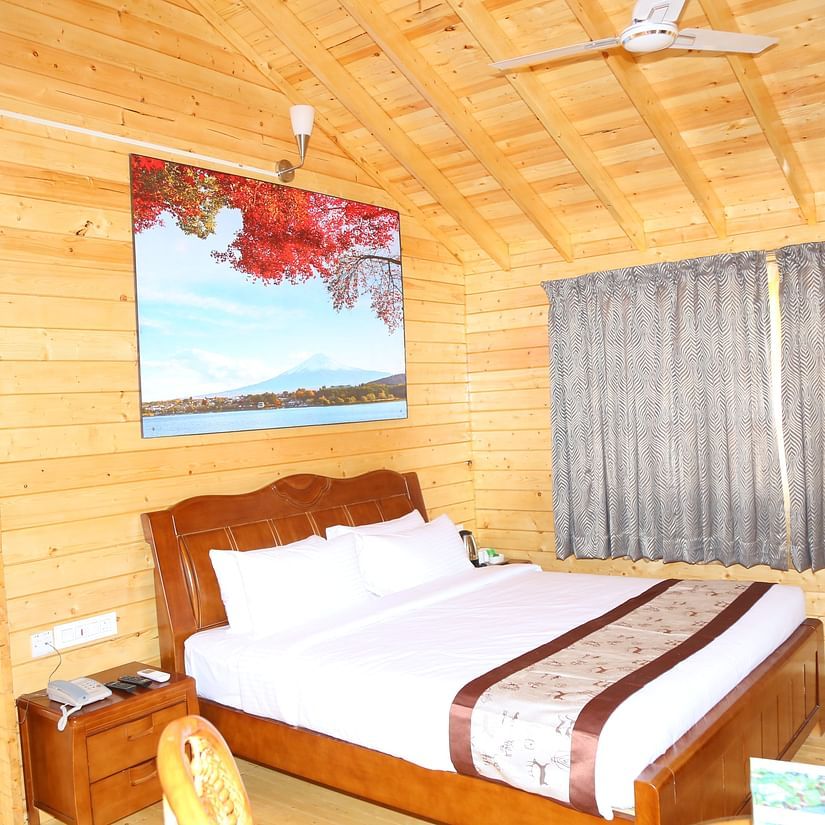 King size bed in a room with wooden interiors, bedside table, curtained windows, ceiling fan at GK Hill View Resort.