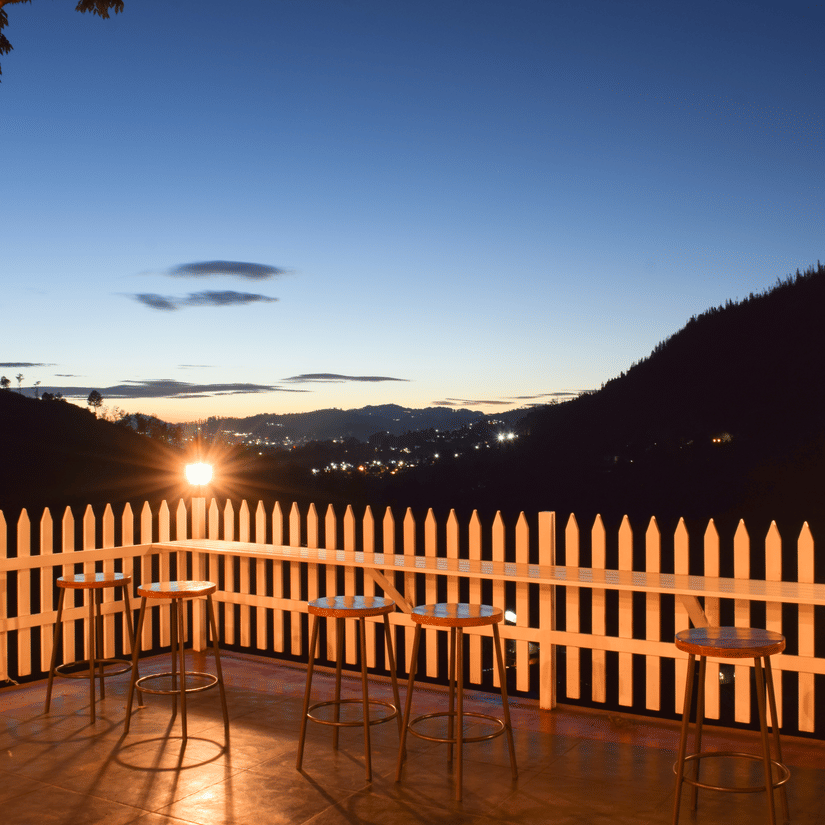 An outdoor patio with a white picket fence at dusk or dawn, featuring tables and chairs, and a distant view of rolling hills under a gradient sky.