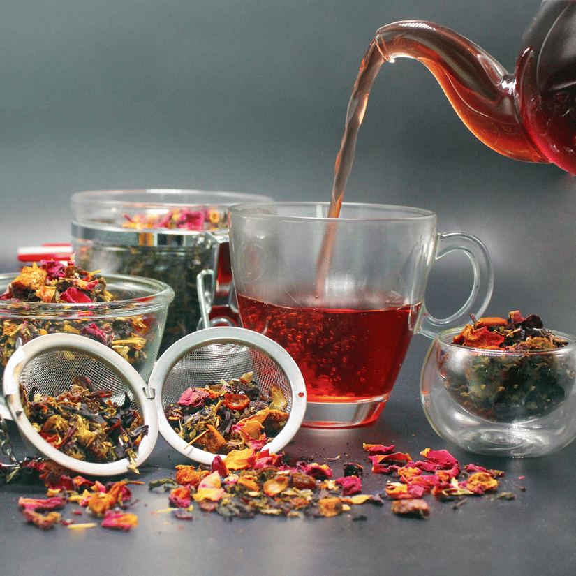 A teapot pours a reddish liquid into a clear mug, surrounded by loose tea leaves, dried flowers, and two metal tea infusers.
