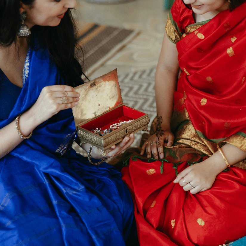 Two people in traditional clothing are seated, with one wearing a blue dress and the other a red sari.