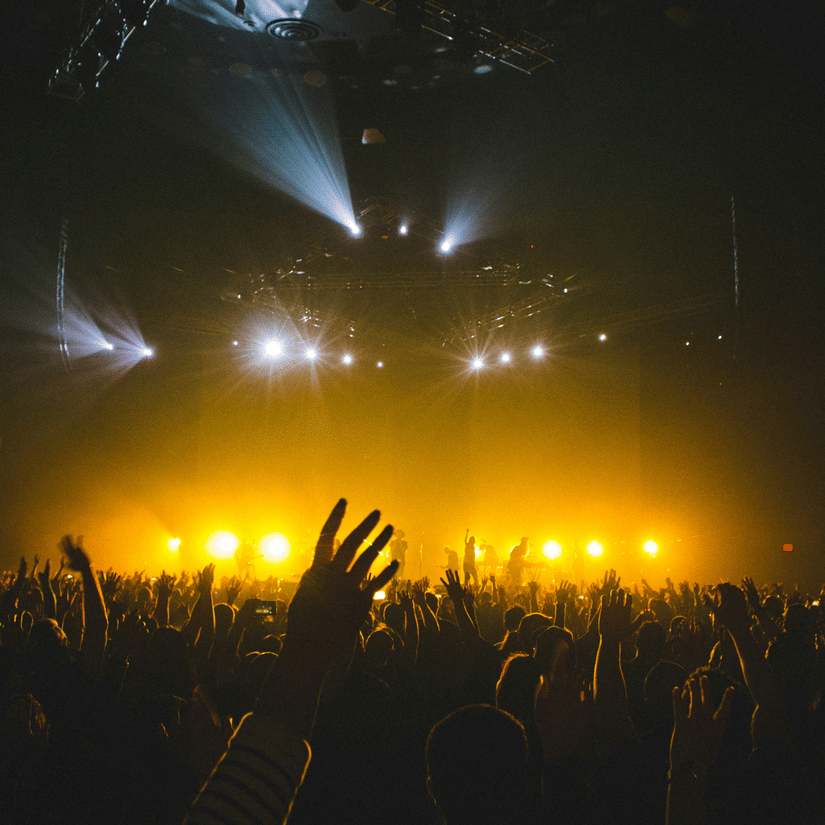 A view from the seating stage of a stage with lights on and a crowd of people dancing to the songs.
