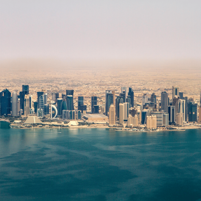 An aerial view of the Doha, Qatar skyline featuring modern skyscrapers along the blue Persian Gulf coast under a hazy sky.