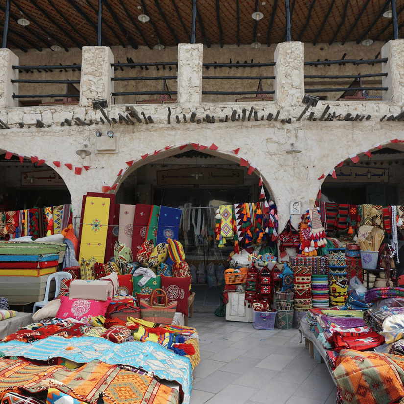 An overview of Souq Waqif market with people selling rugs in a courtyard and an old building in the background.