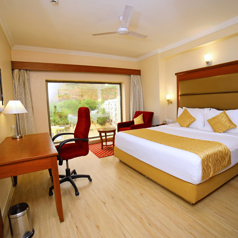 Hill View Room featuring a double bed facing a TV with a lamp on the work desk below, and a sofa next to the window at Raj Park Hotel, offering the best place to stay in Tirupati.
