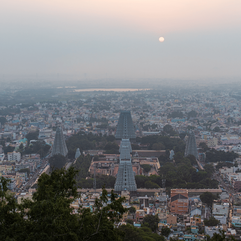 An aerial view of the town of tirupati with numerous temple's gopuram in view and other buildings and trees also in view with the sun setting in the background.