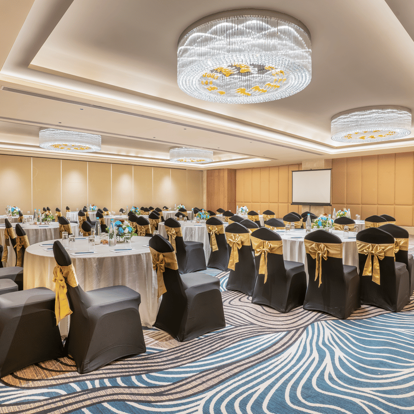 A conference room at Sarovar Premiere Alwar with round tables and chairs with gold sashes and a projector screen.
