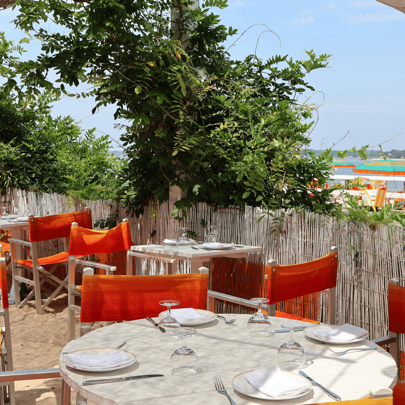 Outdoor beachside restaurant with bright orange director chairs, white tables, and a bamboo fence near the waterfront