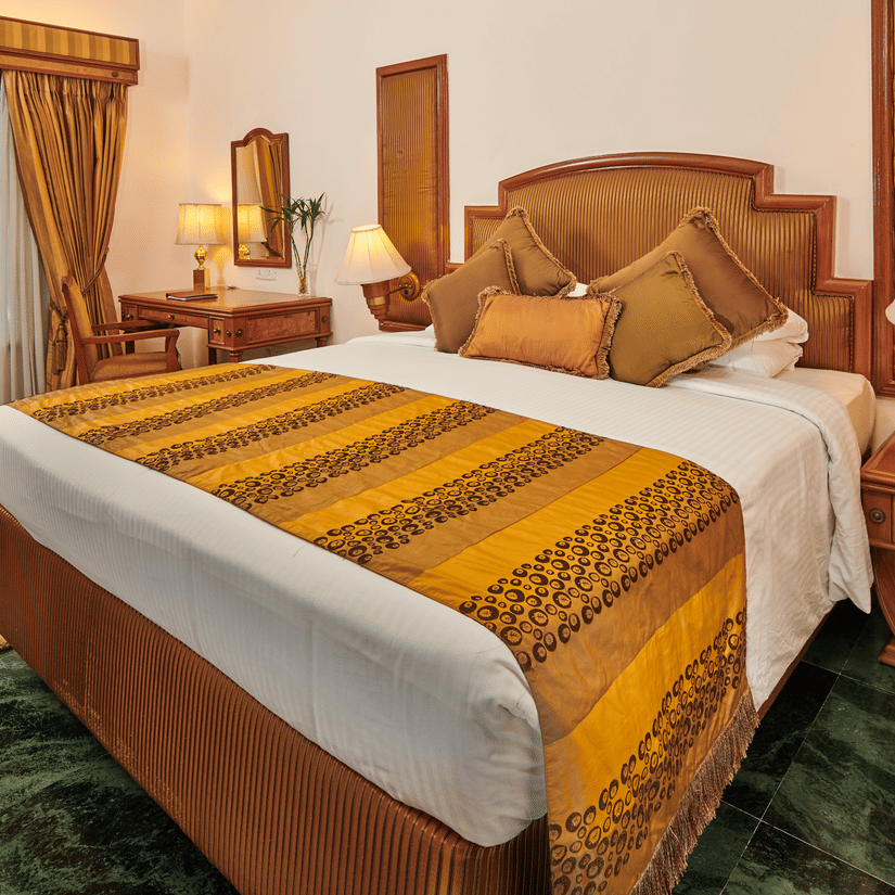 The Ummed Ahmedabad hotel room with a comfortable bed, classic wooden furnishings, and warm ambient lighting.