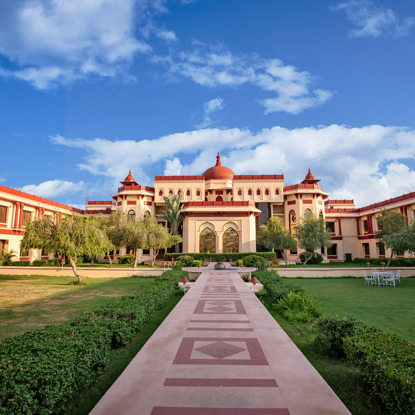Grand exterior view of The Ummed Jodhpur Palace Resort & Spa with a wide pathway, lush green lawns, and clear blue sky.