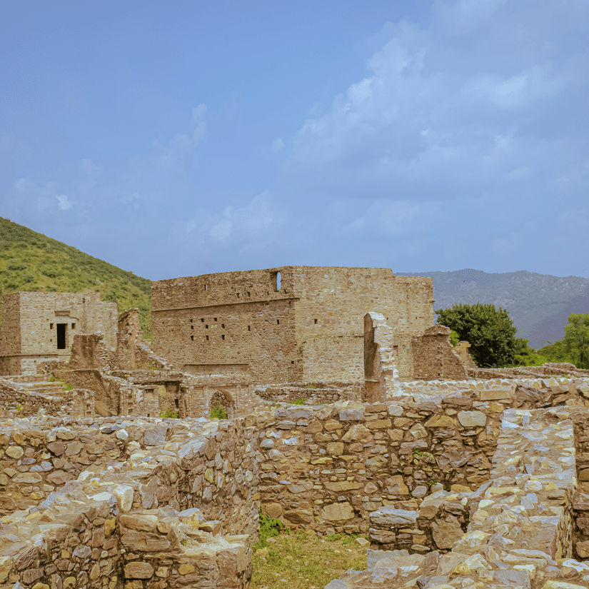 A view from inside Bhangarh Fort of the remains of the stone houses, a mountain on the left hand side and the blue sky in the background.